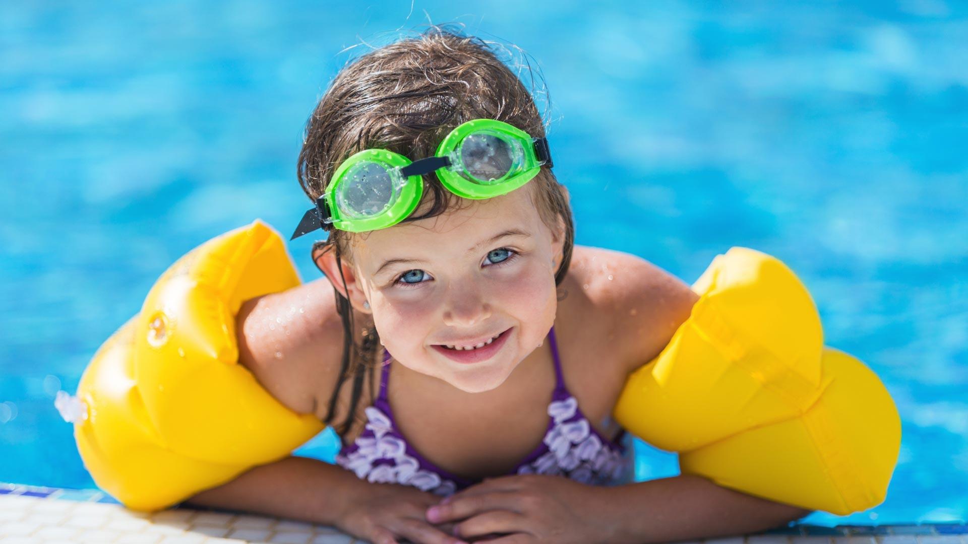 Bambina con braccioli gialli e occhialini verdi in piscina.