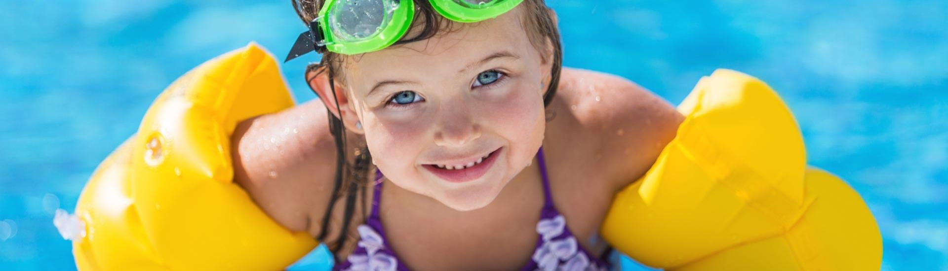 Bambina con braccioli gialli e occhialini verdi in piscina.