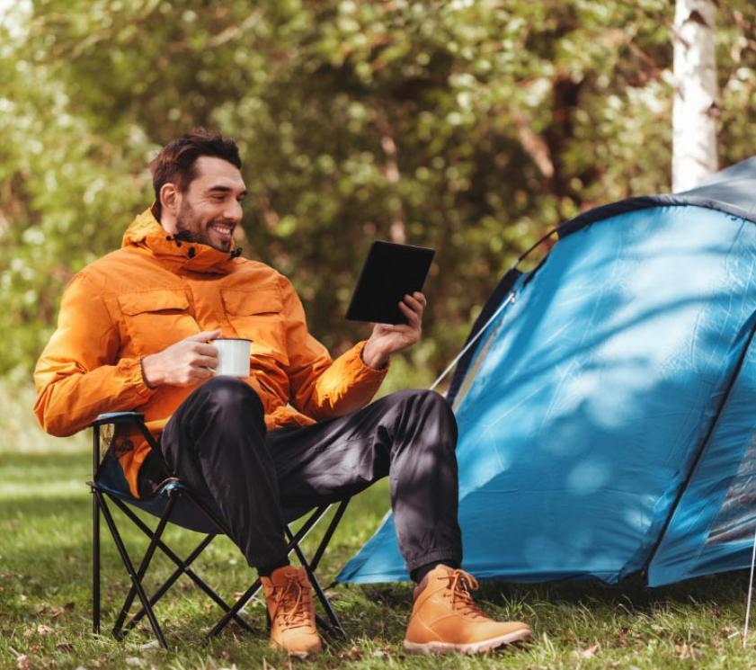 Happy man camping, sitting, with tablet and mug.